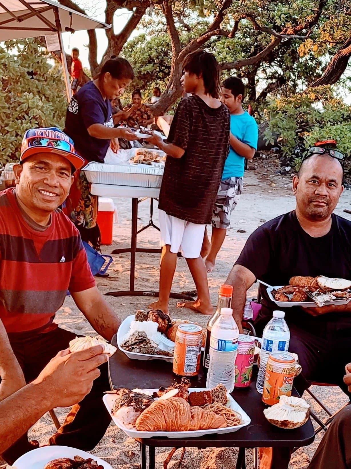 Families eating together at a table outdoors under the trees after a church gathering.
