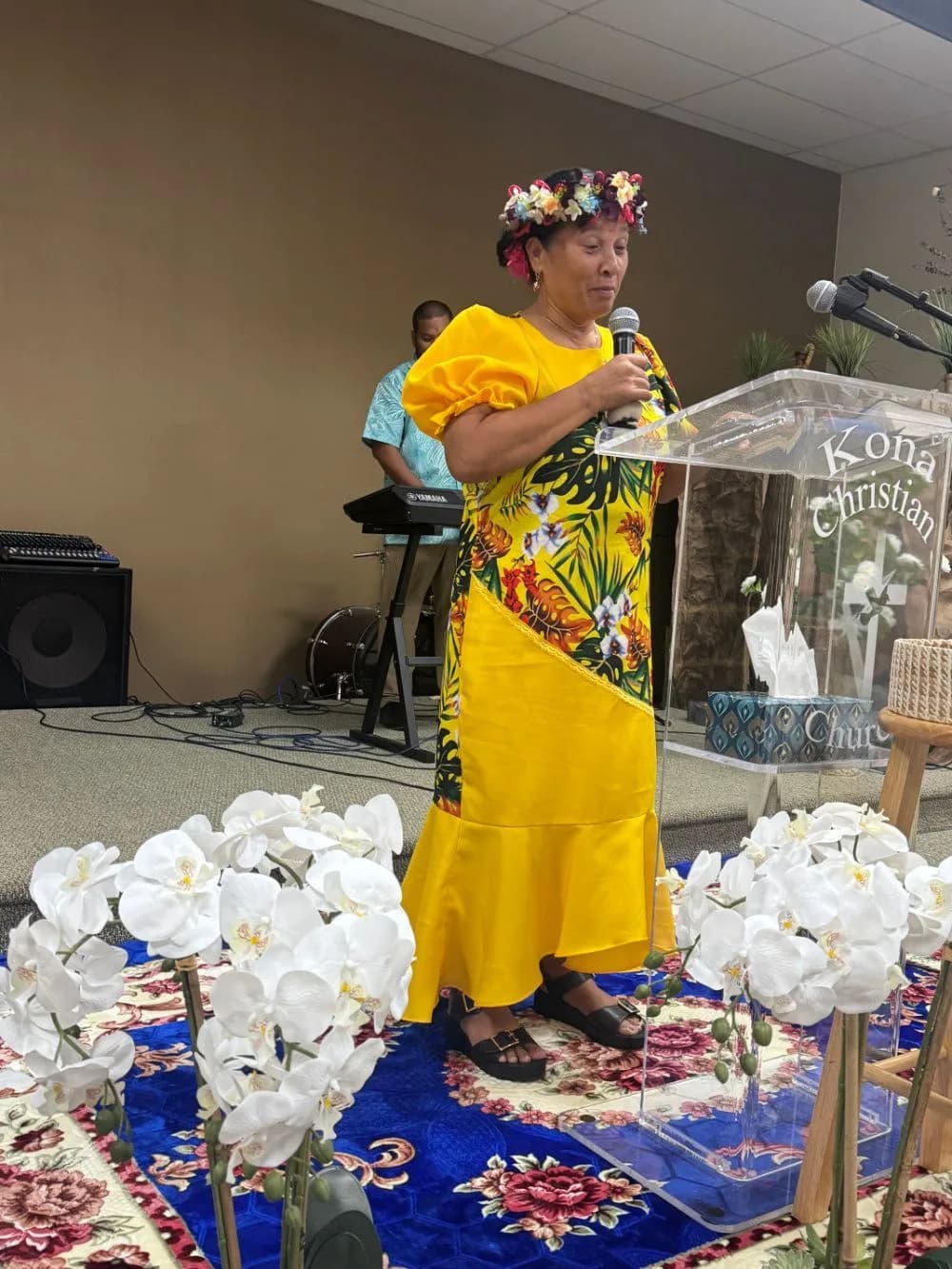 A woman in a flower crown and island dress speaking from the pulpit, with white orchids arranged in front of her.