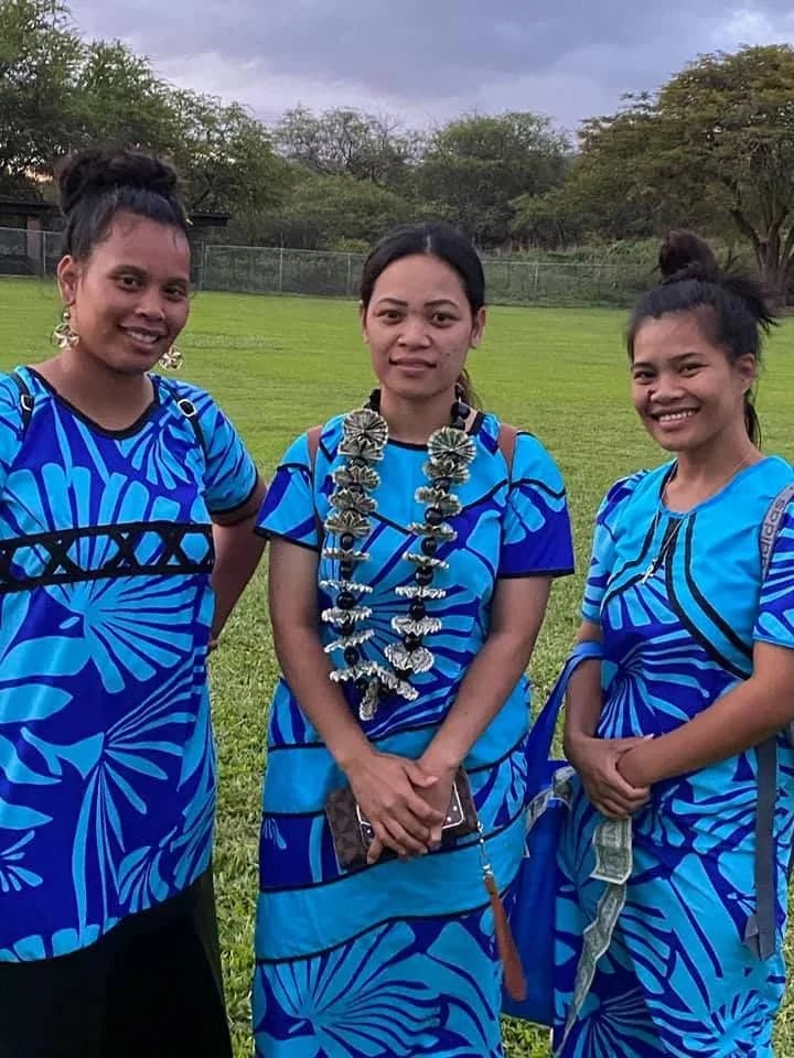 Three young women from the church standing together in blue island dresses and flower leis.